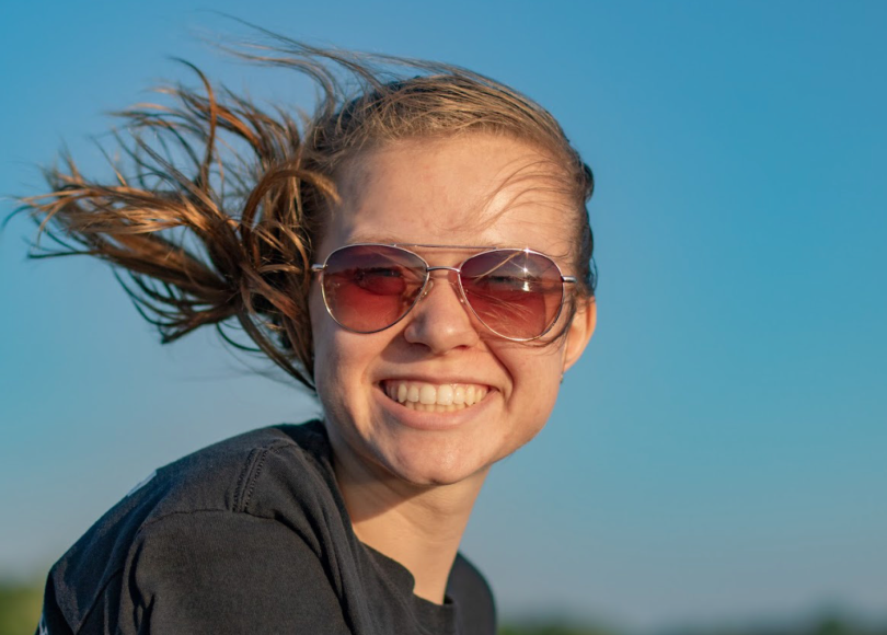 Megan Wolowiec enjoying the summer sun; close-up of smiling woman, wearing sunglasses, long hair blowing in the wind. Photo by Katelyn Wolowiec