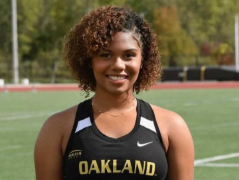 Close-up of smiling young black woman, wearing athletic top that reads Oakland, with athletic field visible behind her